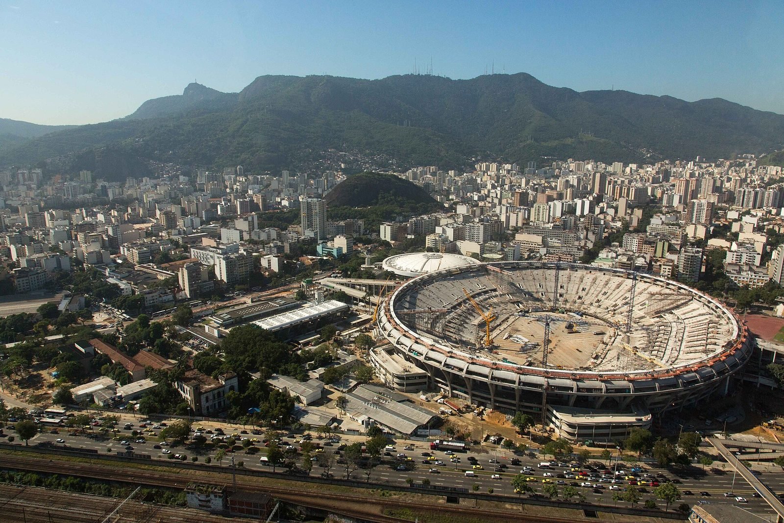 Vista aérea del estadio ovalado sin cubierta, con grúas amarillas en el centro del campo de tierra, rodeado de edificios de la ciudad y cerros verdes al fondo