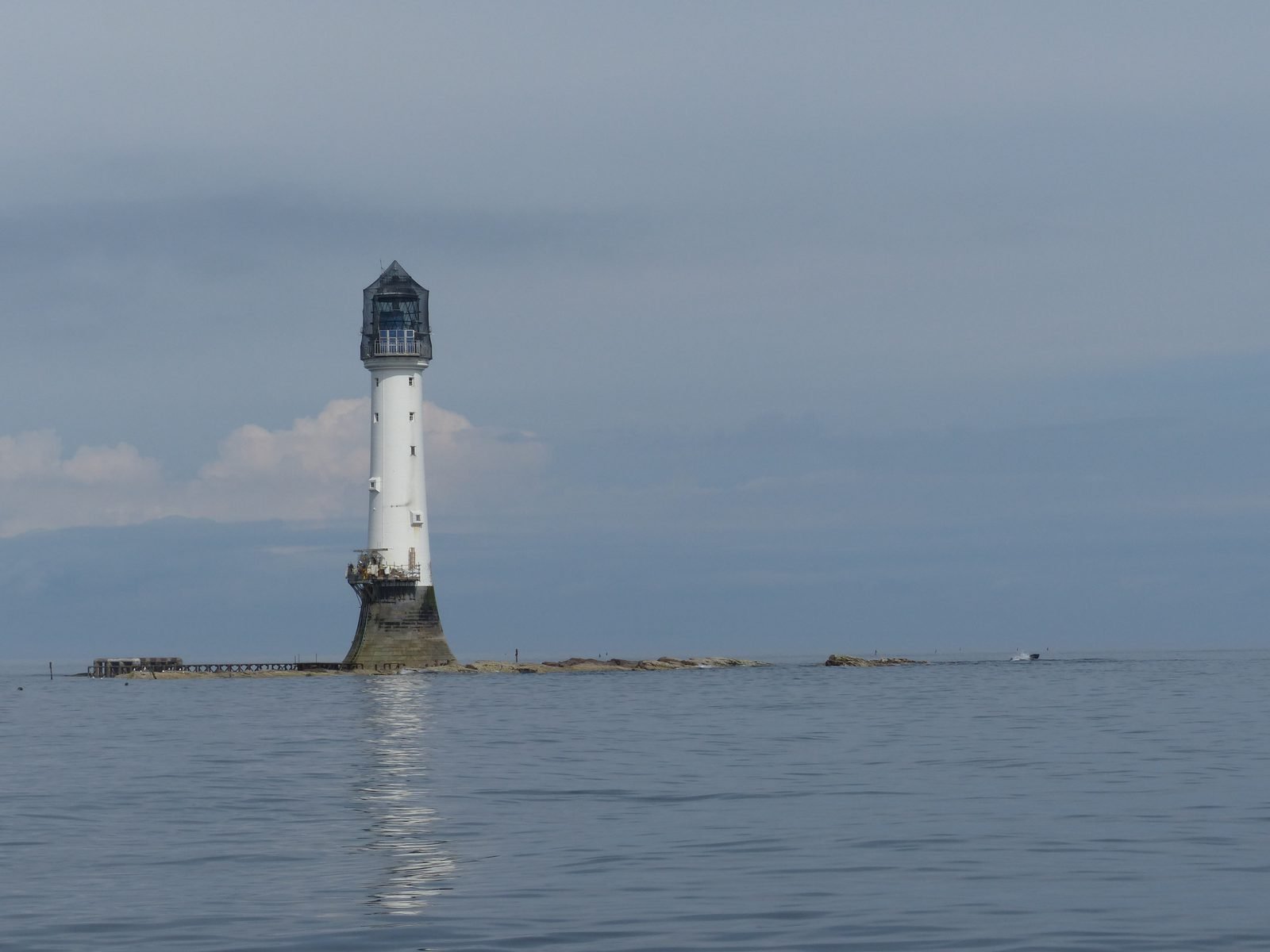 White stone lighthouse with a dark lantern at the top and a wide, dark base, on a low reef almost level with calm water, under a grey-blue sky