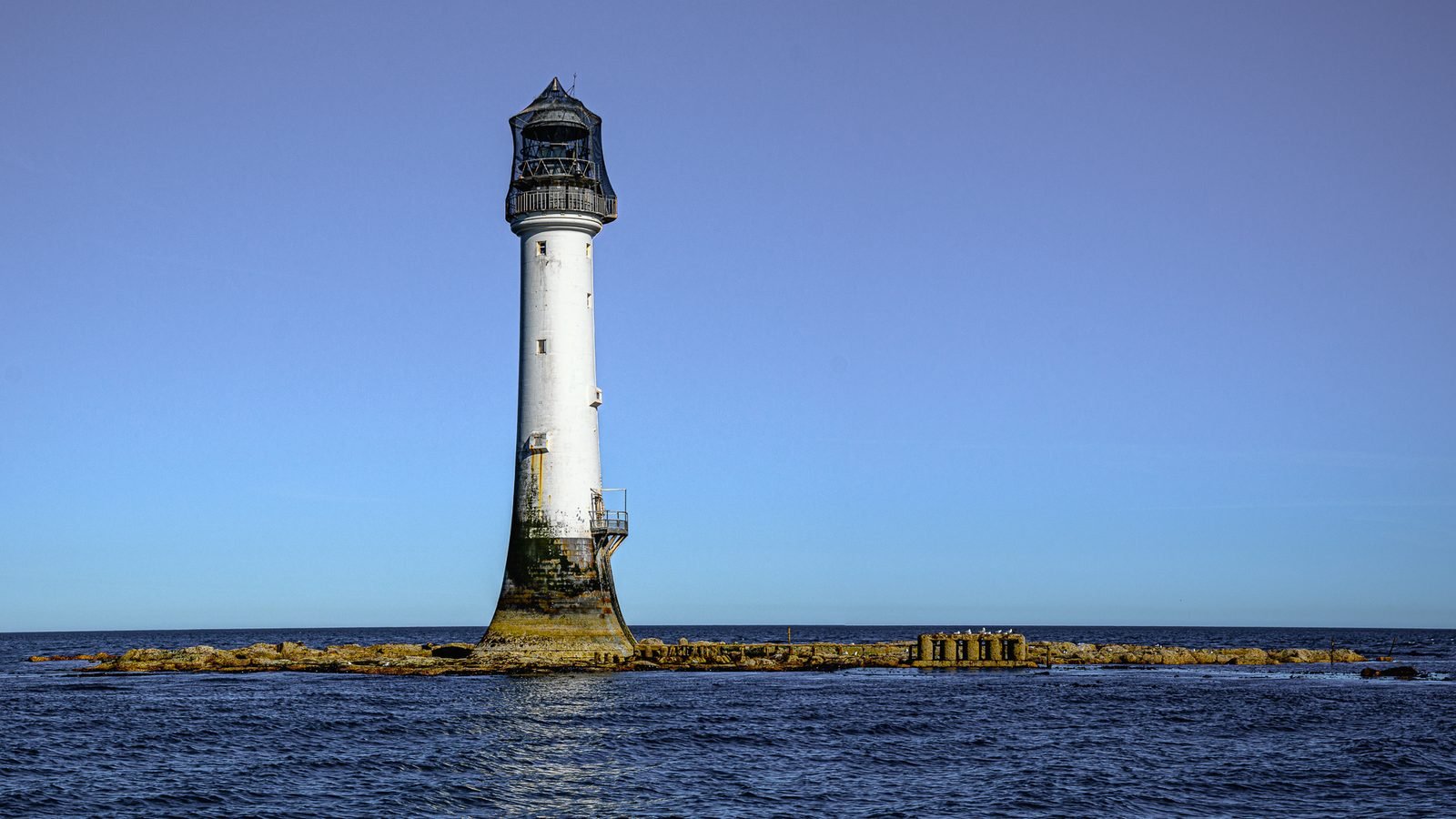 Farol branco com a base de pedra escurecida e úmida sobre um recife baixo, sob céu azul limpo e mar azul-escuro