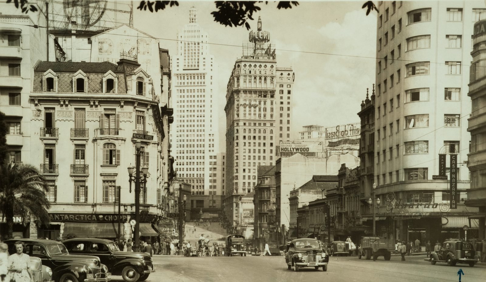 Old sepia postcard: an avenue with 1940s cars, a wide building with a billboard frame on top in the center and, behind it, a taller, stepped white tower