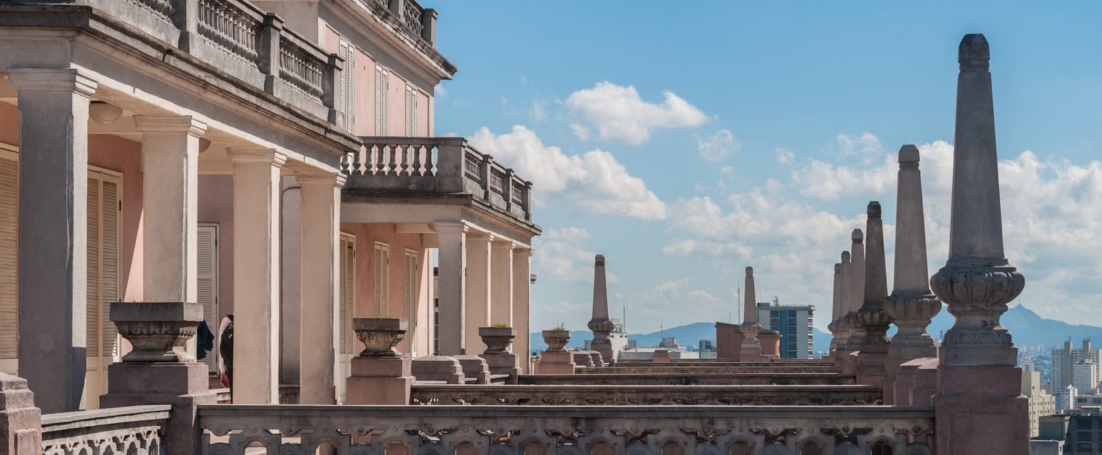 Balconies with columns and balusters on a pink house atop a building, with a row of stone pinnacles and the city and mountains behind under a blue sky