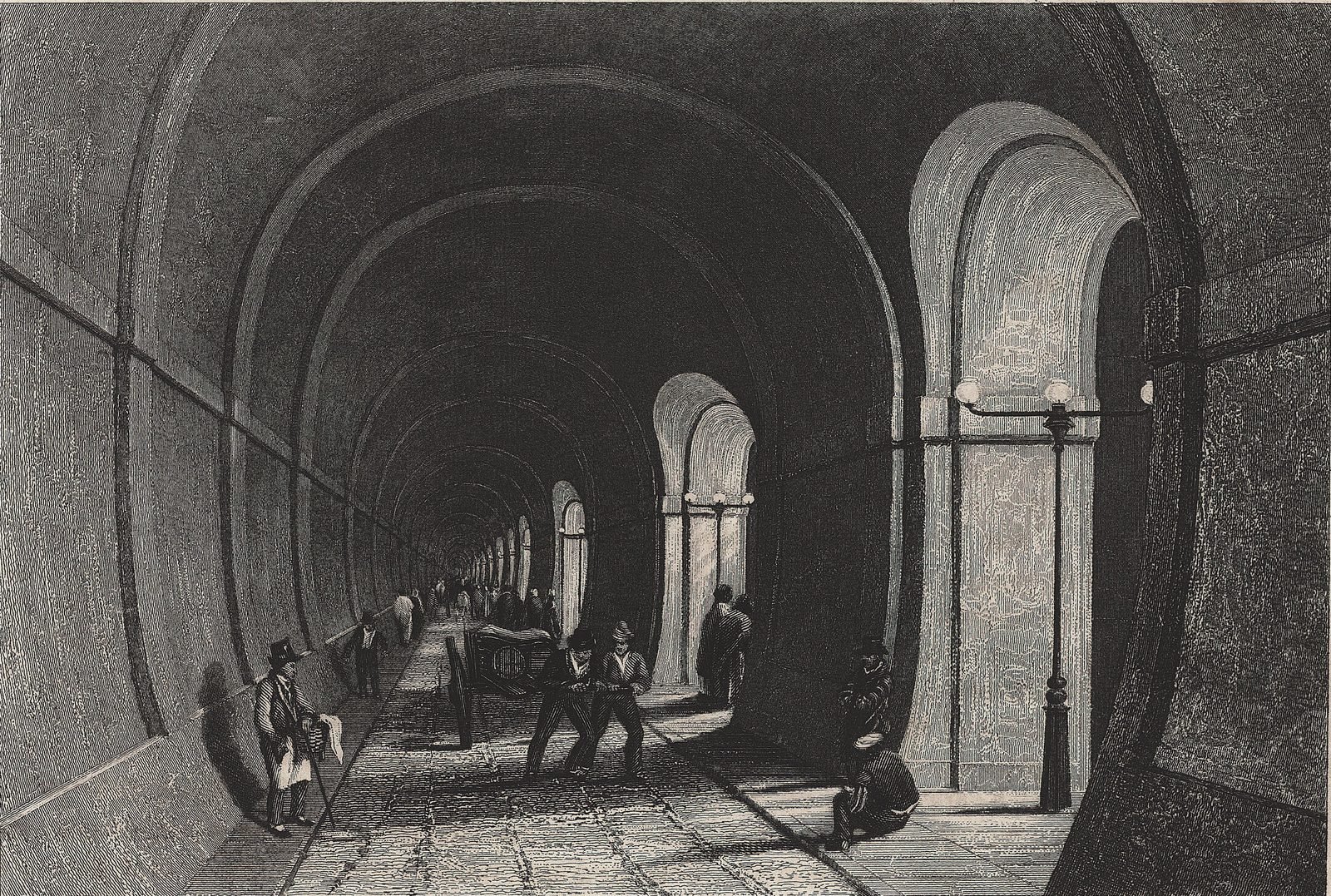 Old black-and-white engraving: an arched brick gallery receding into the distance, with side archways lit by lamps, pedestrians in top hats and two men pulling a cart on the stone floor