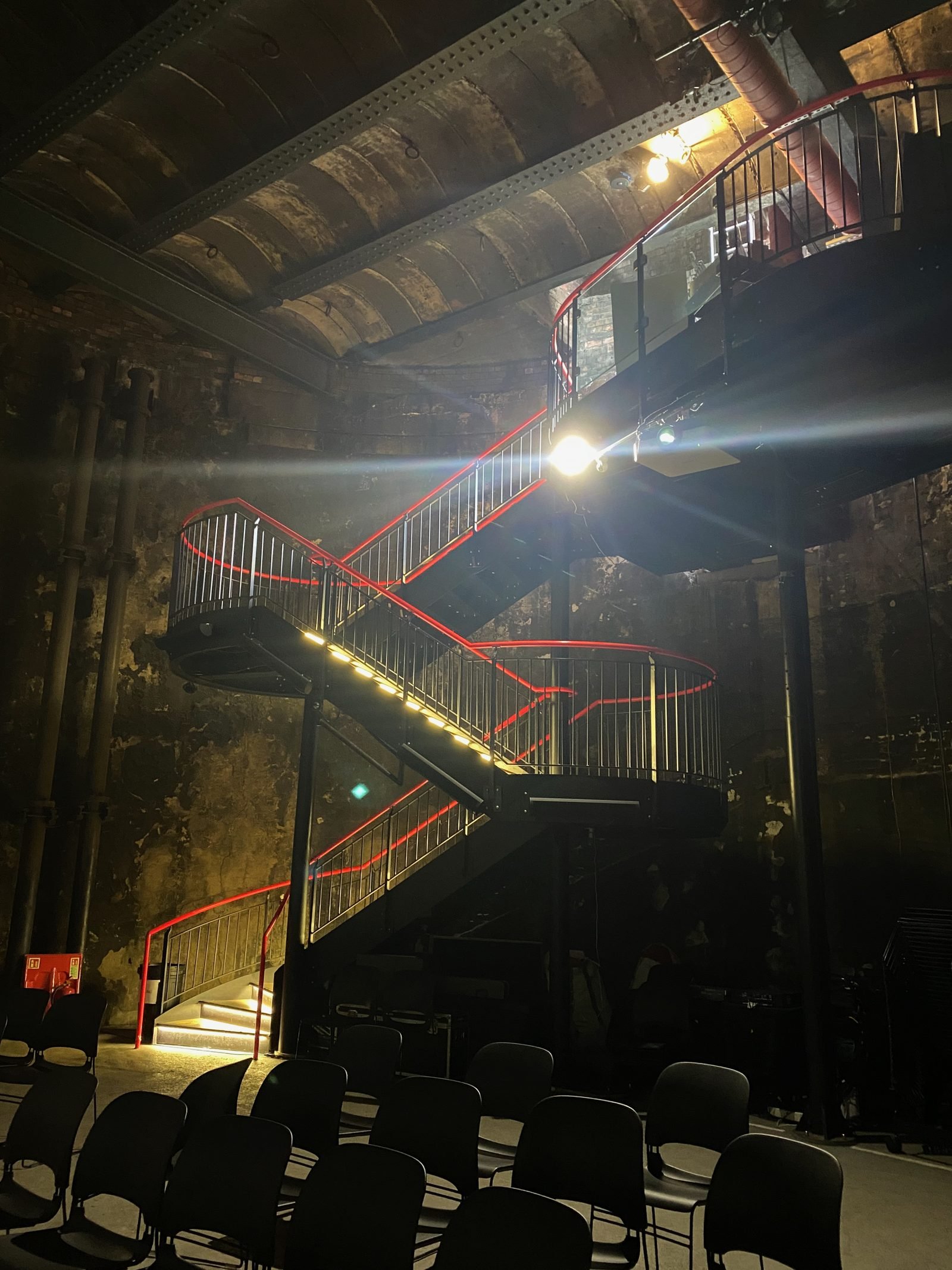 Dark interior of a circular shaft of blackened brick, with a zigzag metal staircase outlined in red light and chairs on the floor