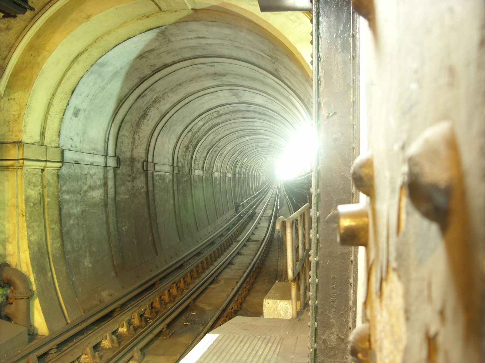 Inside an arched tunnel with train tracks running toward a bright light in the distance; on the left, the original columns and arches of painted brick