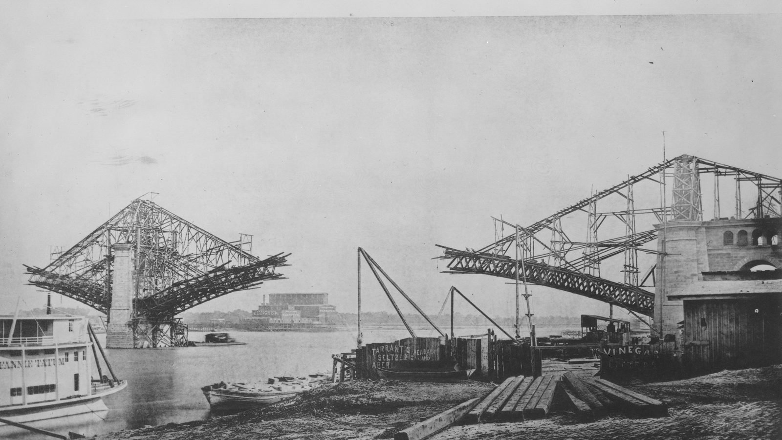 Fotografía antigua: en la orilla empedrada, un medio arco de acero sale del estribo de mampostería sujeto con cables a una torre; en el río, una pila con dos medios arcos y un vapor amarrado