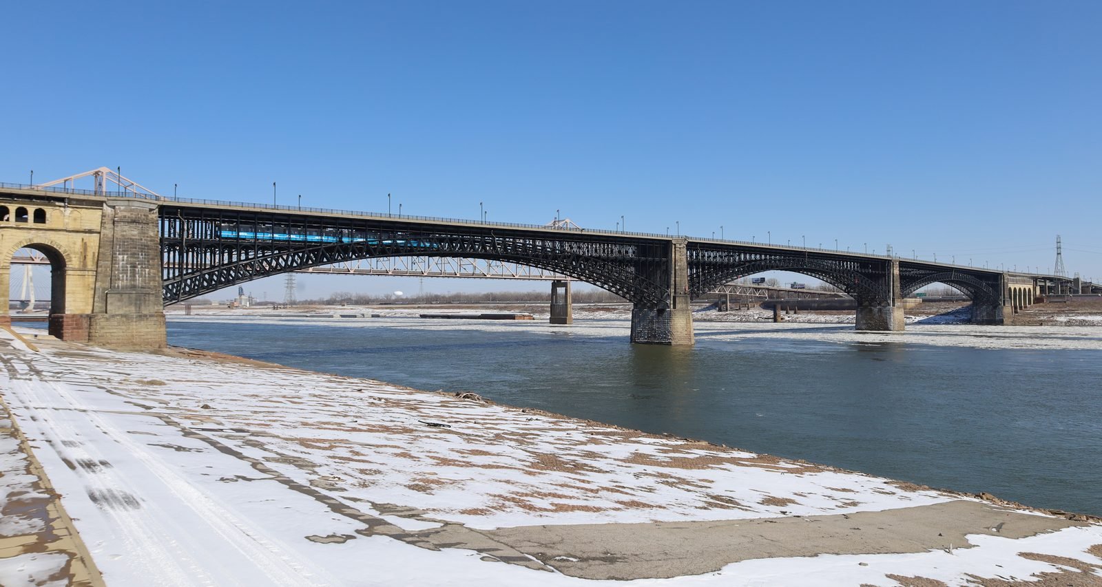Bridge of three dark steel arches over a wide river, with stone piers, a blue train crossing on the lower deck, a clear sky and a snow-covered bank in the foreground