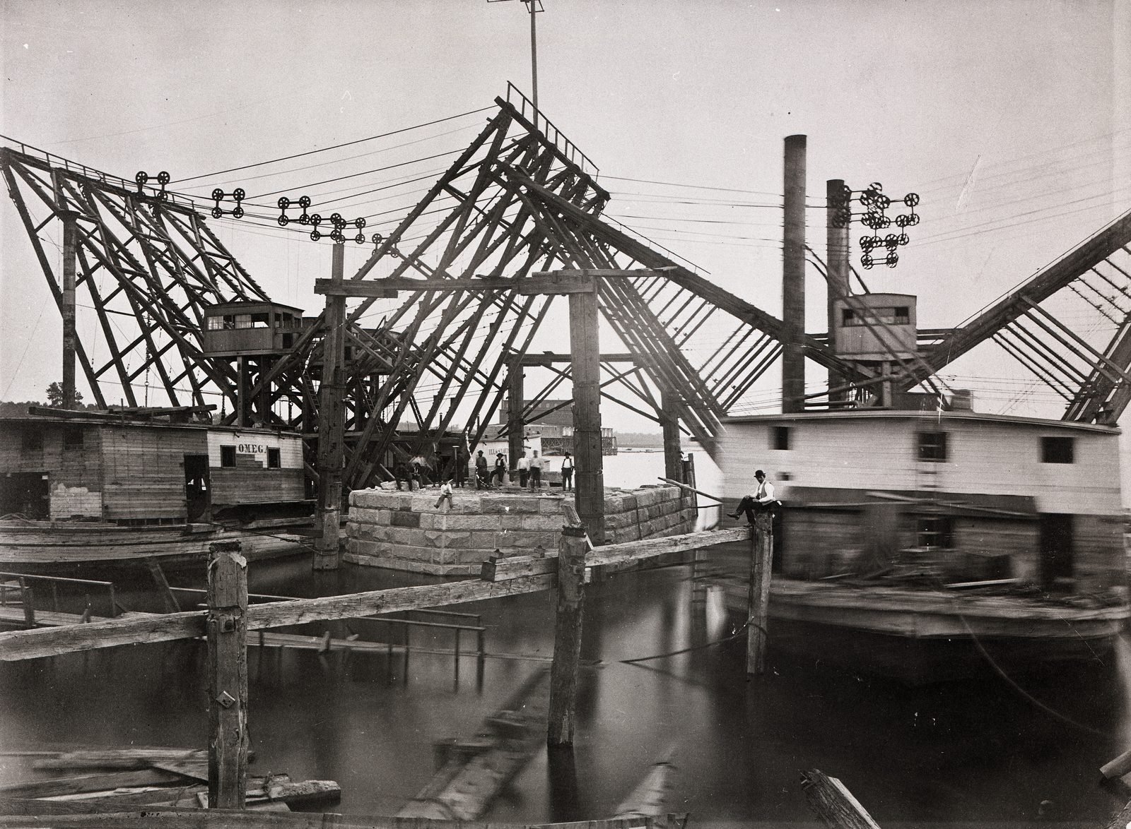 Fotografía antigua en blanco y negro: un bloque de mampostería que emerge del agua entre barcos taller de madera, bajo grandes armazones inclinados de madera con poleas, chimeneas y hombres sobre las piedras