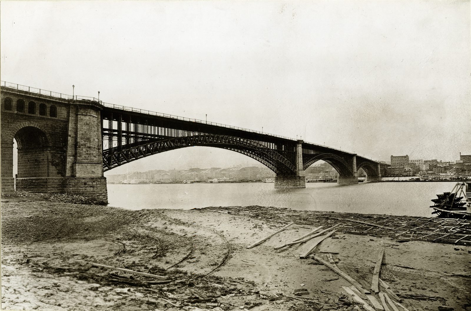 Fotografía antigua en sepia: el puente de tres arcos visto desde una playa de arena con tablones sueltos, el estribo de piedra con arcadas a la izquierda y la ciudad de St. Louis al fondo