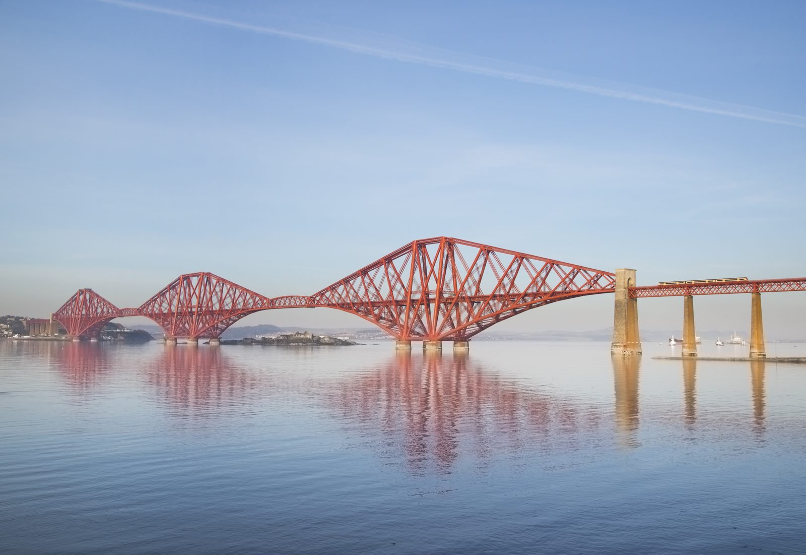 Red steel bridge with three large diamond-shaped structures over calm water that reflects them, a stone pier on the right and a train crossing the viaduct under a blue sky