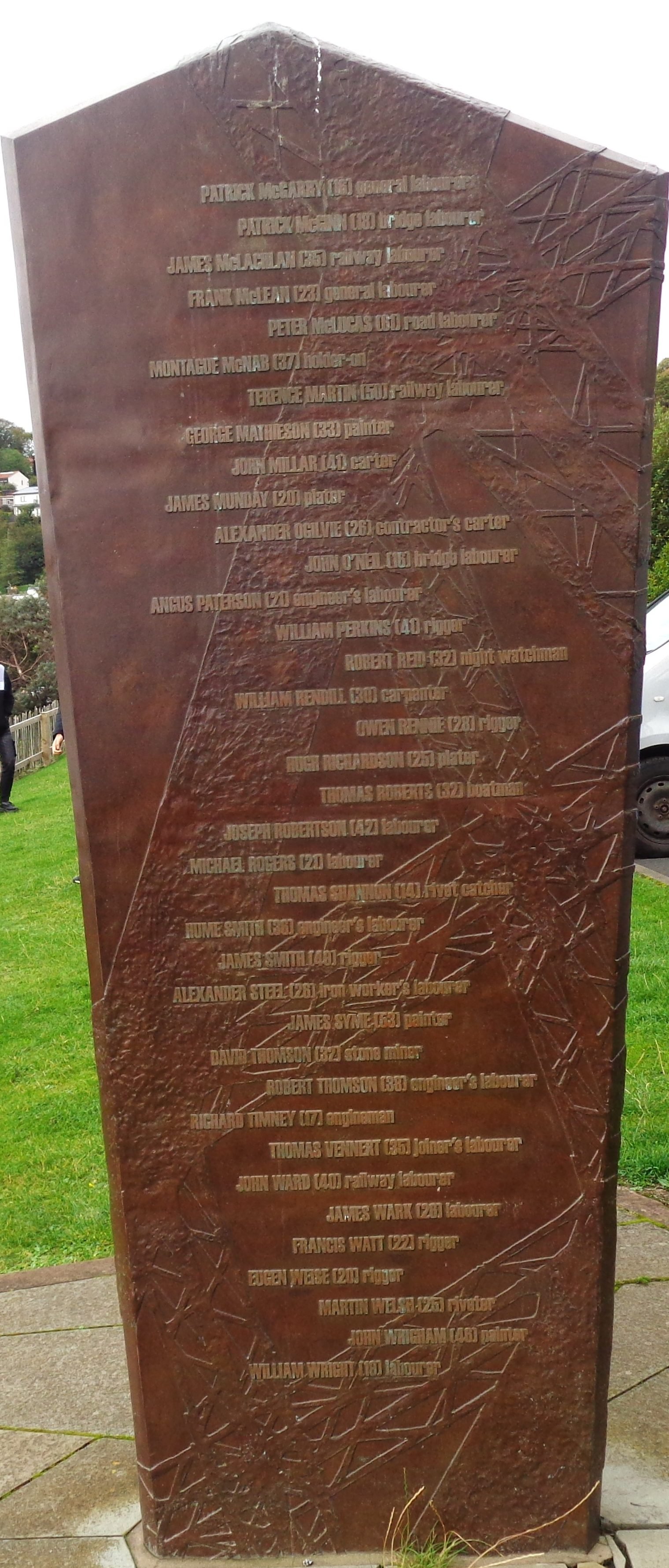 Monumento alto de bronze em forma de lâmina, com colunas de nomes gravados, sobre um gramado, com casas e árvores ao fundo