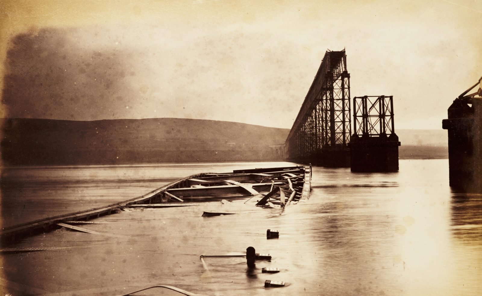 Fotografia antiga em sépia: vigas de ferro tombadas e retorcidas saindo da água de um rio largo, com uma fileira de pilares de ponte ao fundo terminando num vão vazio