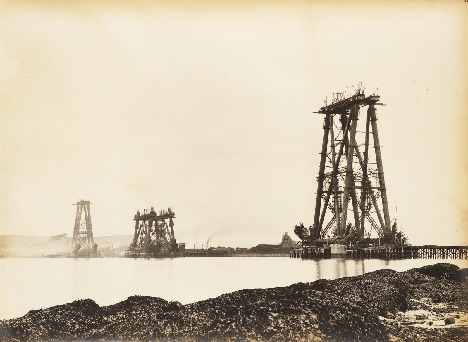 Fotografia antiga em sépia: três torres de treliça de aço em construção erguidas sobre a água de um estuário, ainda sem os braços, vistas de uma praia com rochas cobertas de algas