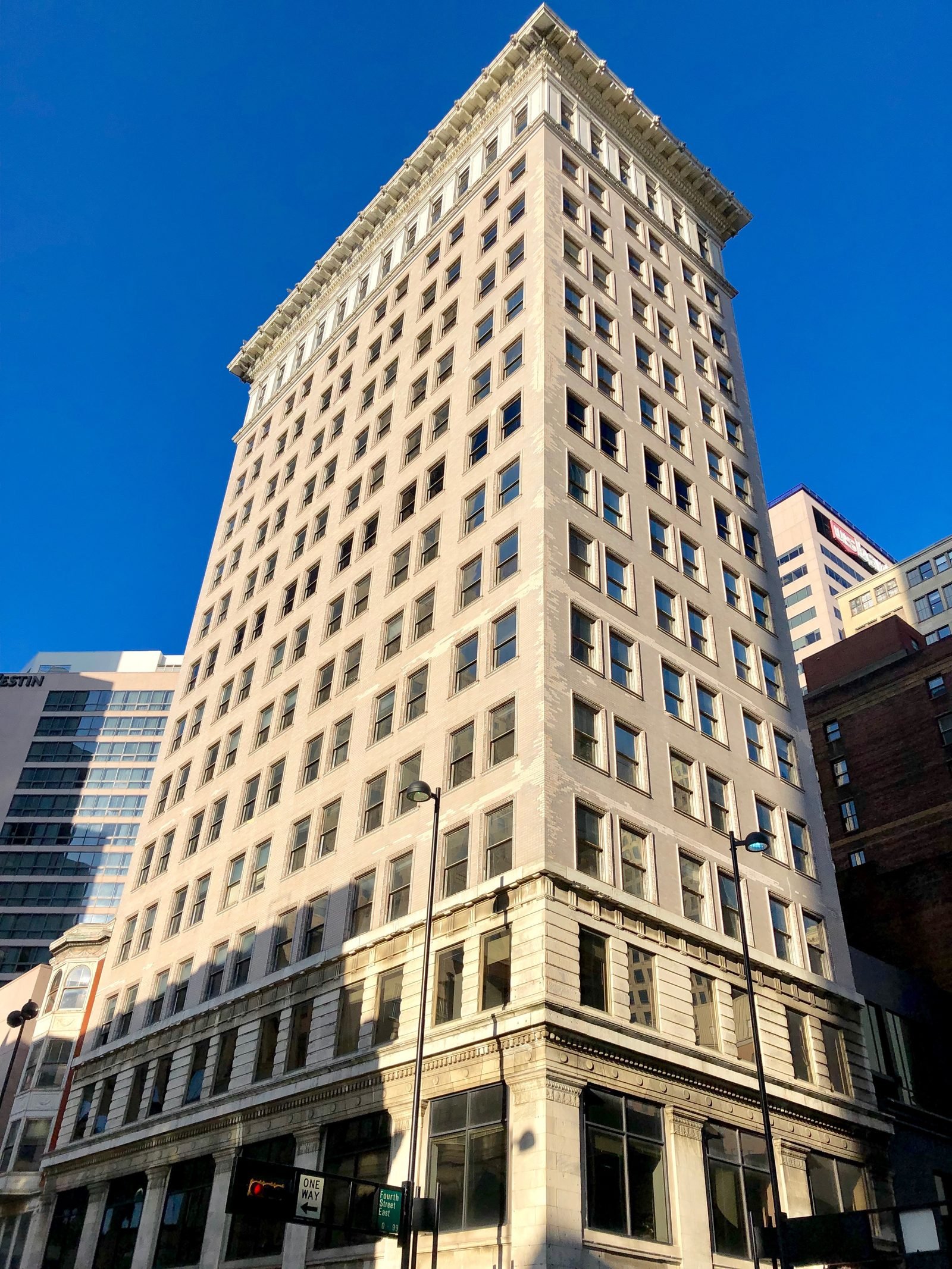 Tall, narrow building with a light-colored facade, seen from below at the corner, with a stone base, regular rows of windows and a projecting cornice at the top, against a blue sky