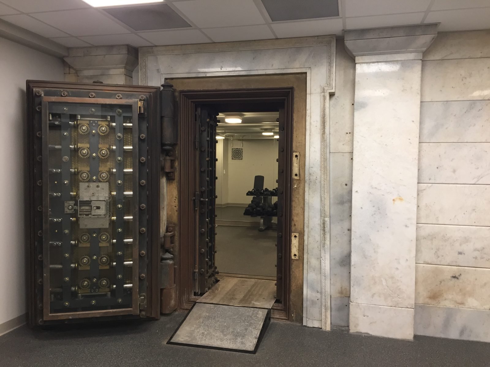 Rectangular bank vault door, open, full of bolts and gears, in a marble wall; inside, a small gym with dumbbells