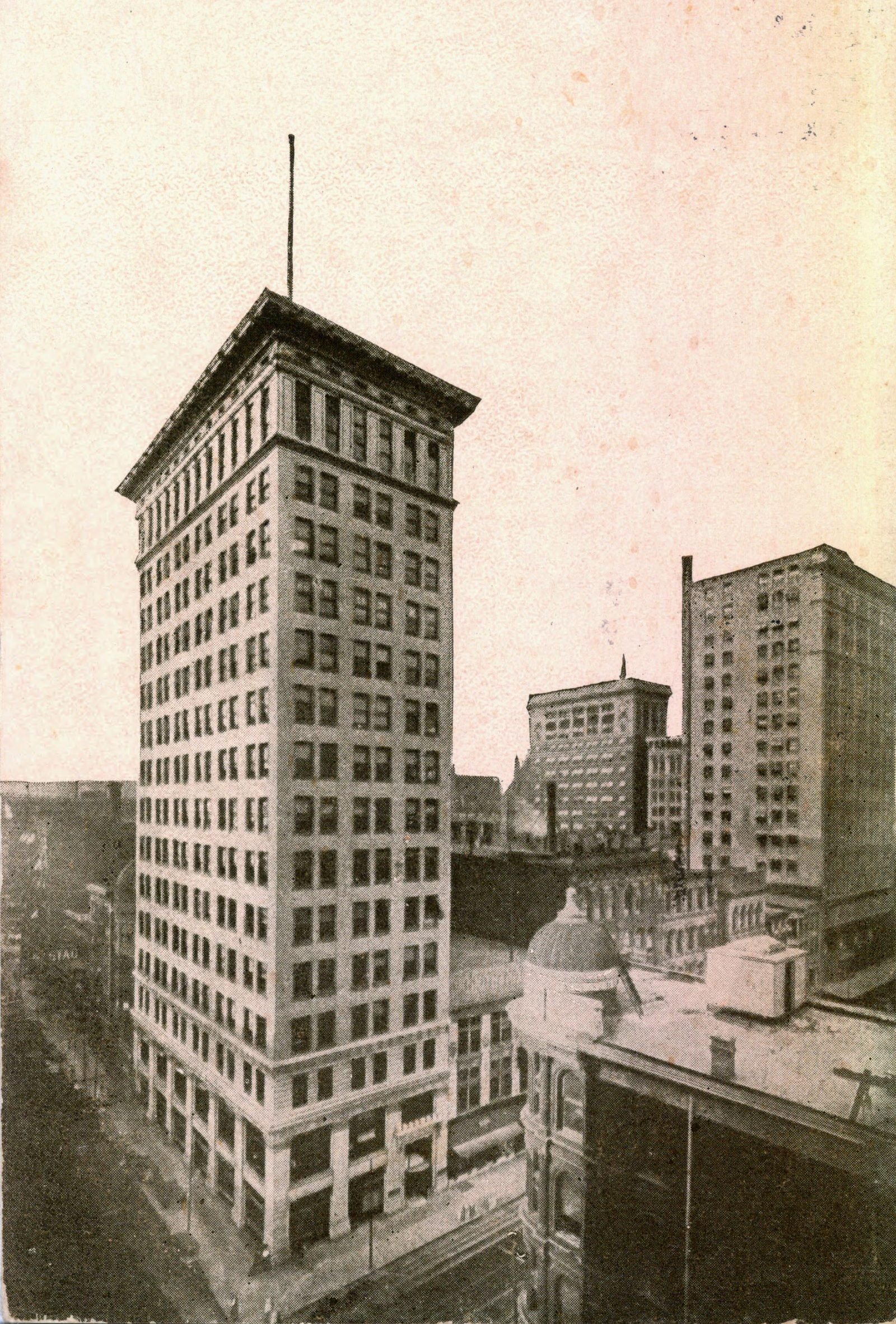 Old sepia postcard: a tall corner building seen from above, with a flagpole on top, the street far below and lower buildings around it