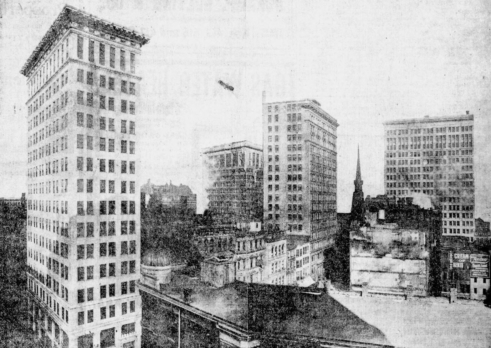 Black-and-white newspaper photograph: on the left, a tall, light-colored building dominates the scene; on the right, three more distant tall buildings, a church spire and a small airship in the sky