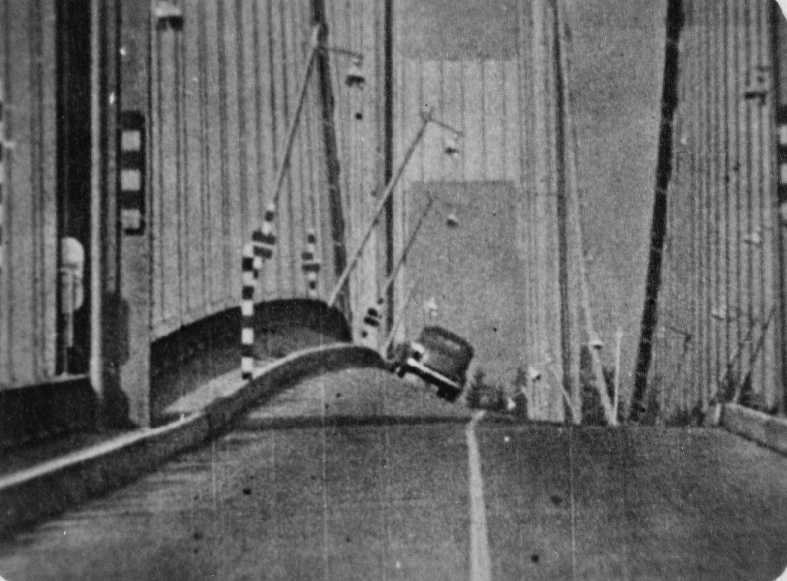 Old black-and-white film frame: the bridge roadway tilted and twisted, with a car stopped and tilted in the middle, bent lampposts and the vertical suspenders on both sides