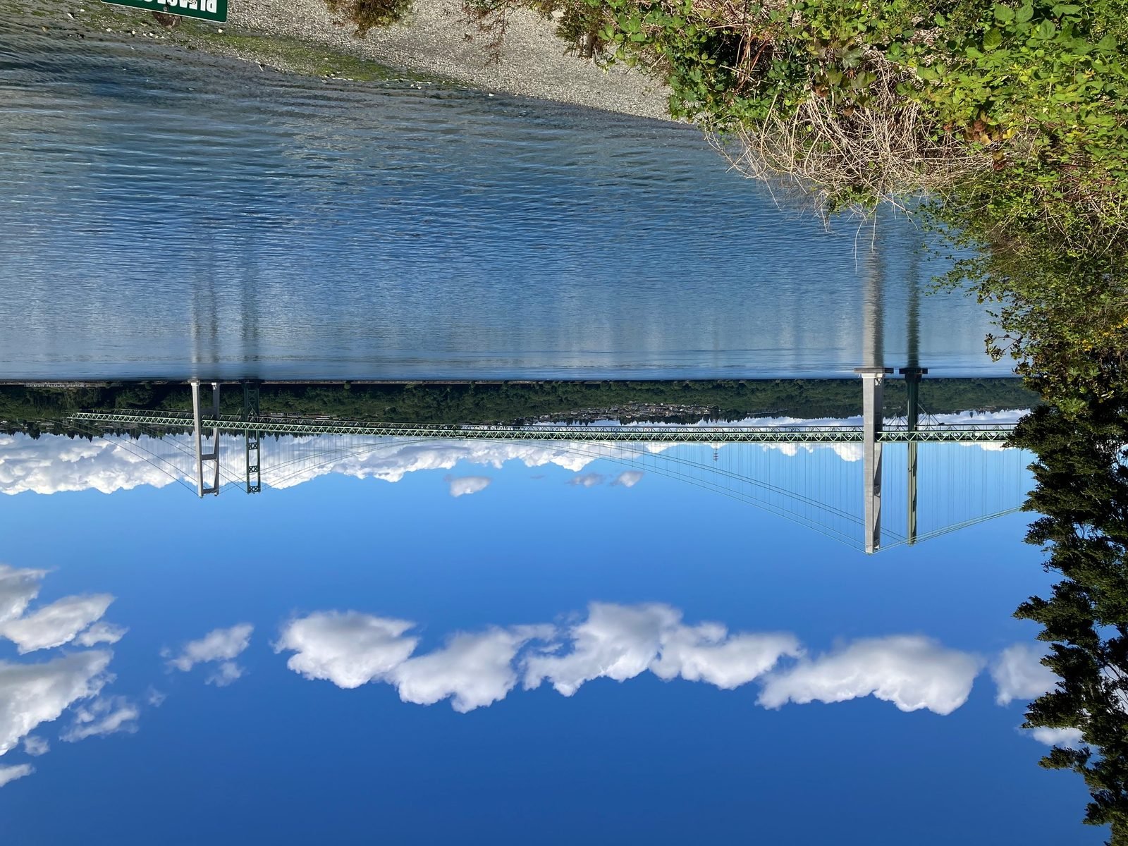 Two parallel suspension bridges over a strait of blue water, under a sky with white clouds, seen from a beach with vegetation; the nearer one has light concrete towers and the one behind, green steel towers