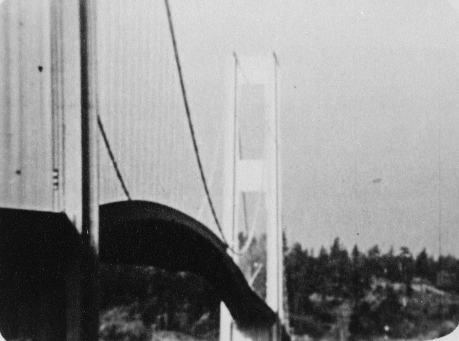 Old black-and-white film frame: the bridge roadway arched like a dark wave, seen from near the tower, with the main cable curving down and the other tower in the background