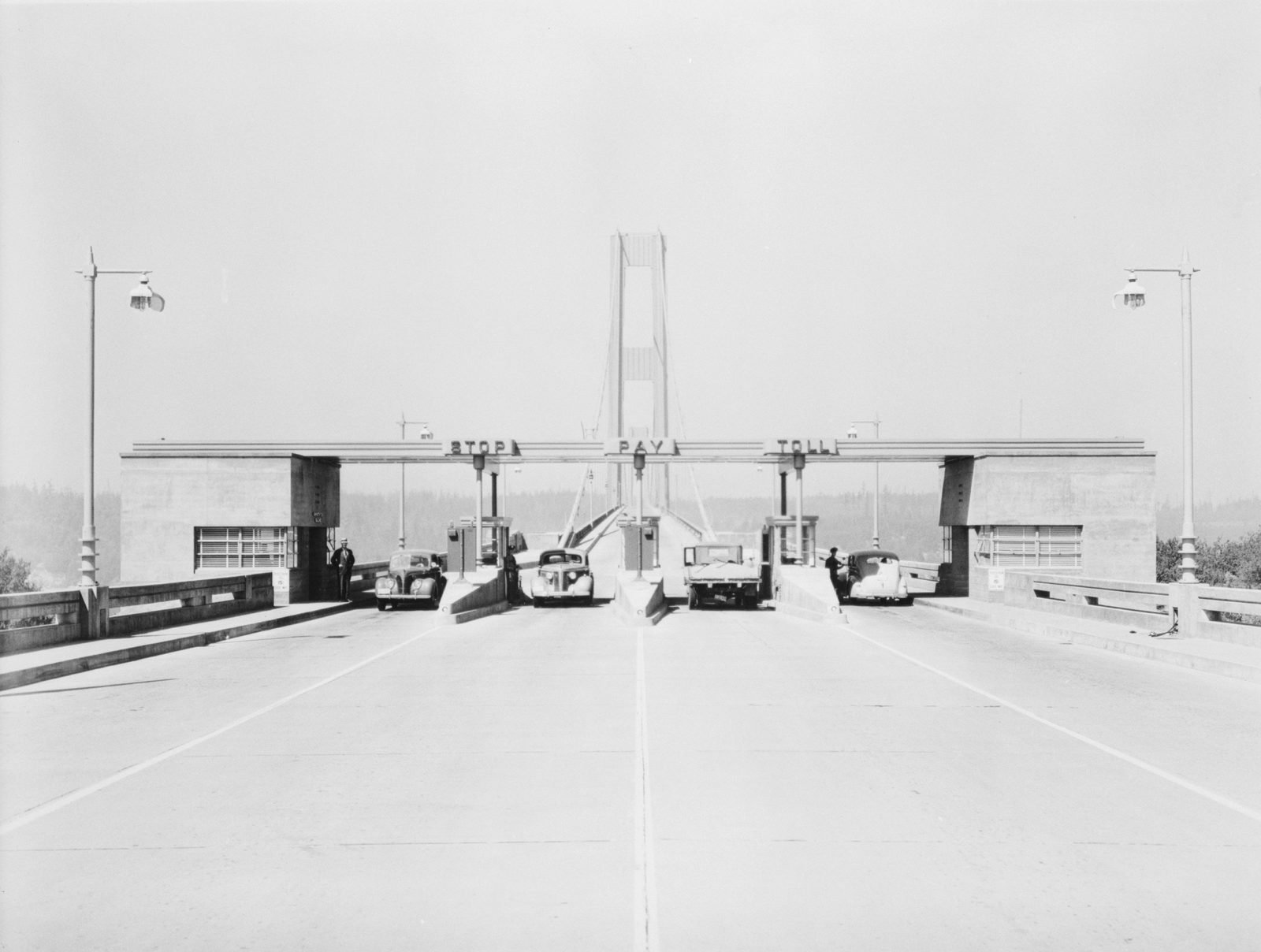 Old photograph: a toll plaza with four booths under a canopy bearing the words Stop, Pay, Toll, a few cars from the 1940s stopped and the bridge's slender tower in the background, in the mist