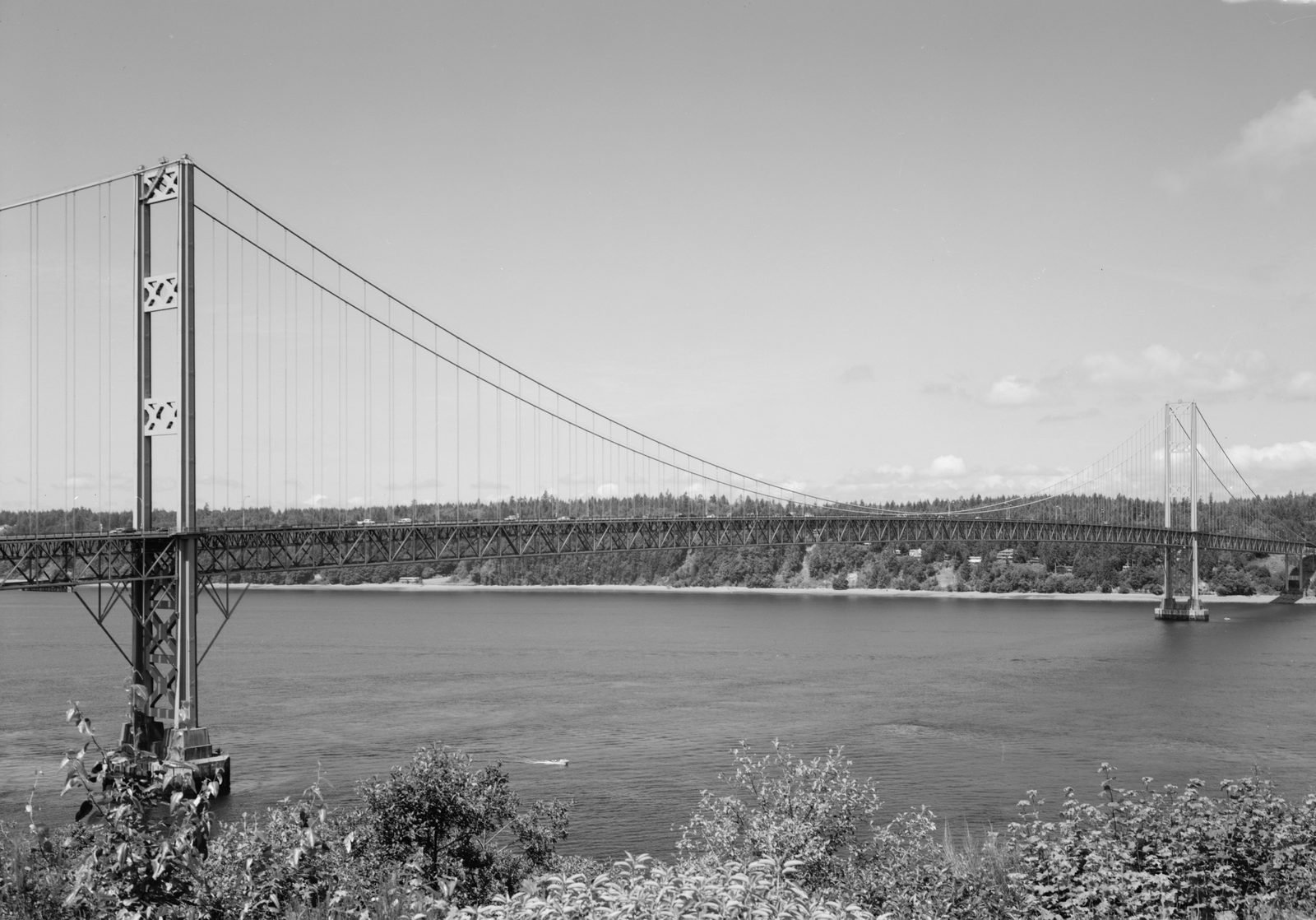 Black-and-white photograph of a long suspension bridge over calm water, with a lattice tower on the left in the foreground, the roadway resting on a deep truss and the second tower far off on the right