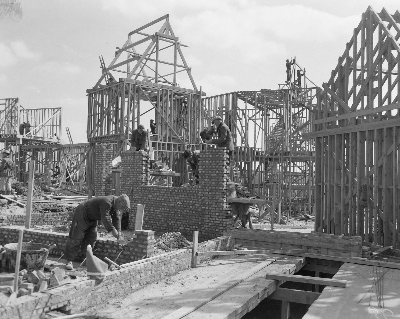 Fotografia em preto e branco: operários levantando paredes de tijolo entre estruturas de madeira de casas em construção, num terreno de obra cheio de material