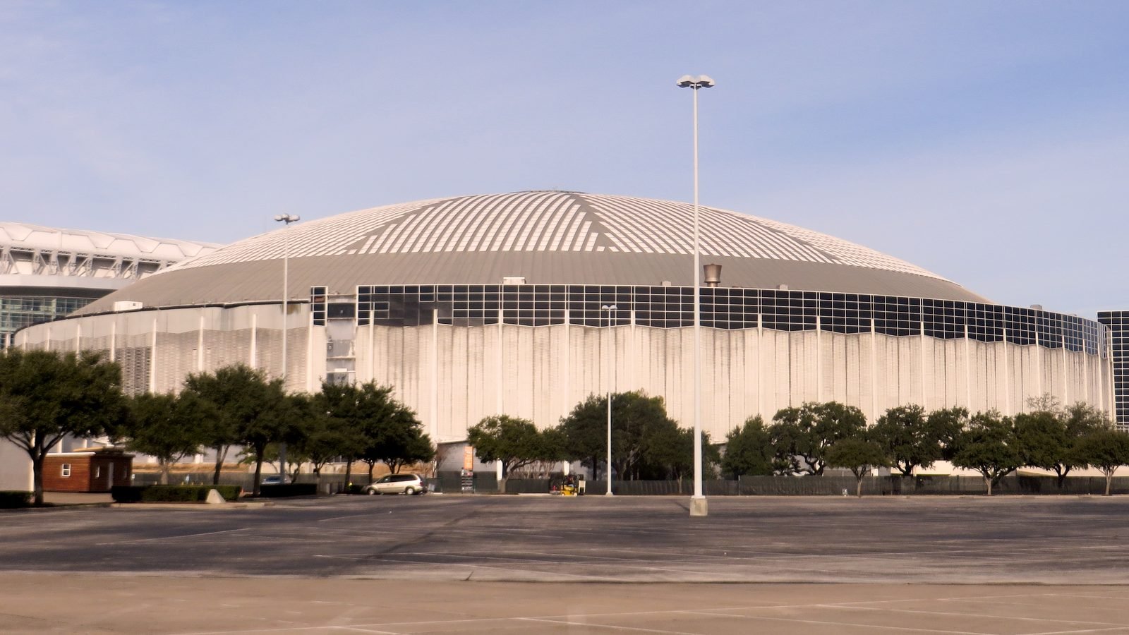 O estádio fechado visto do estacionamento, com a cúpula listrada, a parede de concreto manchada e árvores na frente