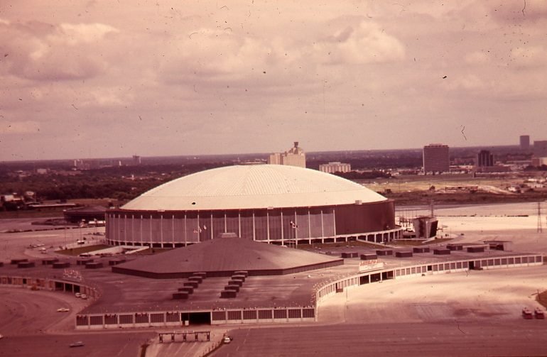 Fotografia colorida desbotada: a cúpula branca do estádio sobre a parede circular escura, com um pavilhão de teto baixo e estacionamento em primeiro plano