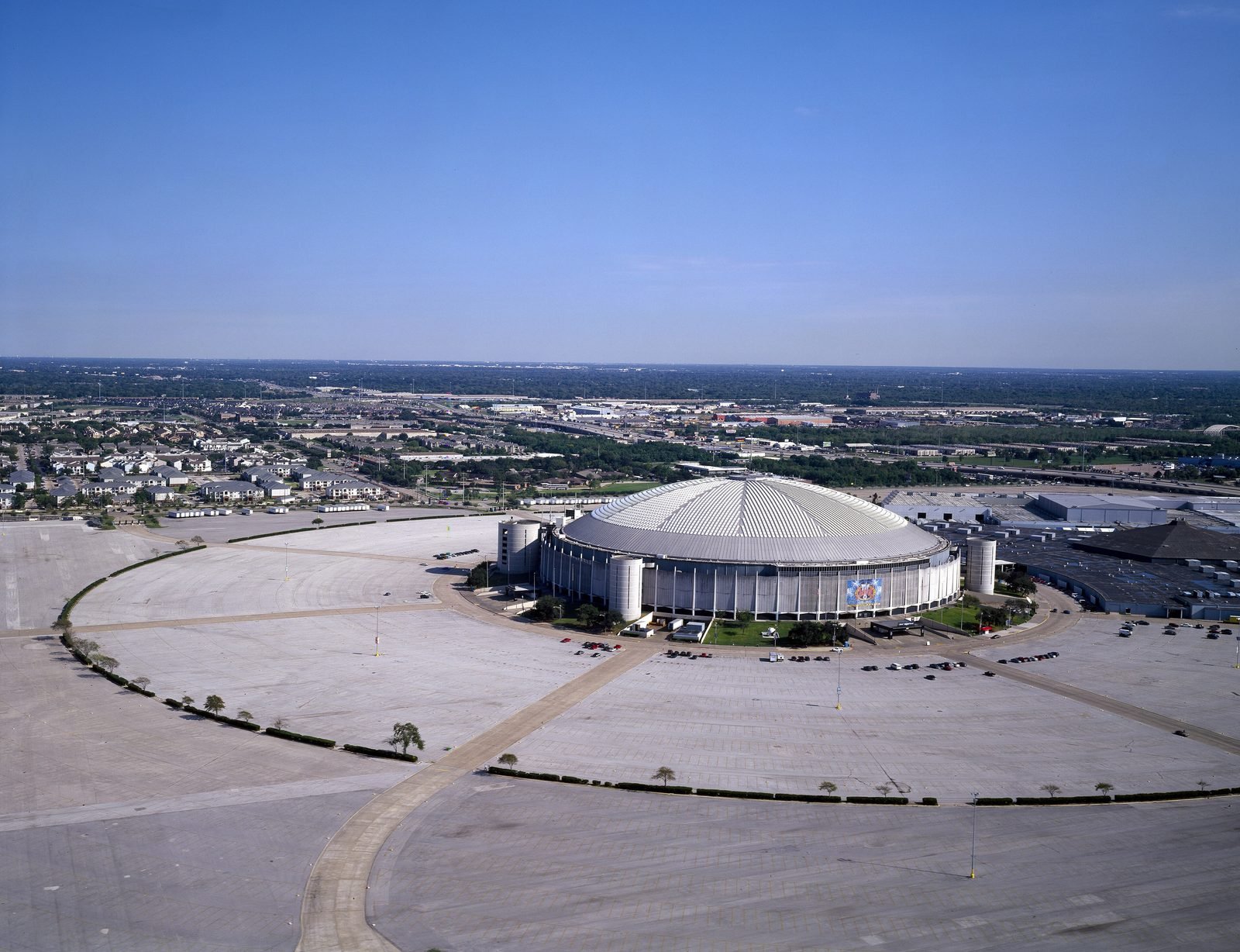 Aerial view of a round stadium with a low, light-colored dome in the middle of a huge empty parking lot, with Houston neighborhoods and highways in the background under a blue sky