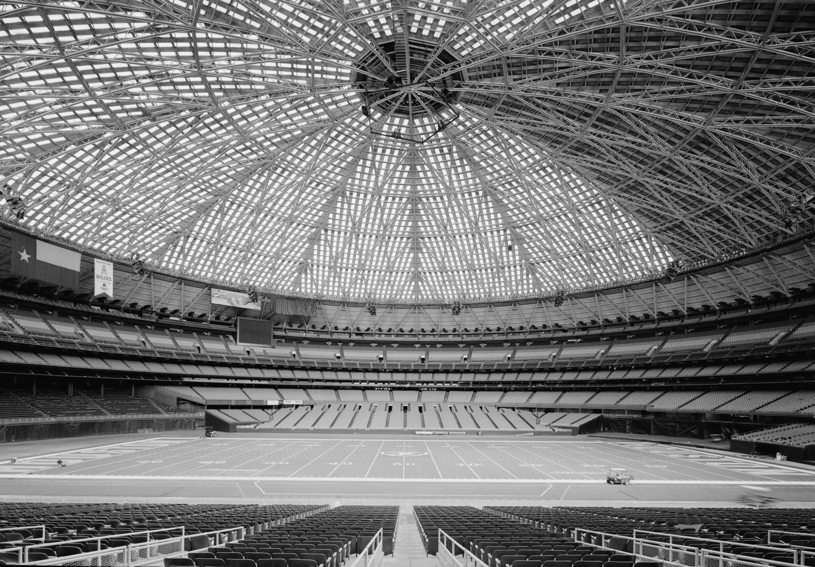Fotografia em preto e branco do interior: a cúpula inteira vista de dentro, uma malha de vigas de aço em losango irradiando de uma lanterna central, sobre arquibancadas vazias e um campo de futebol americano