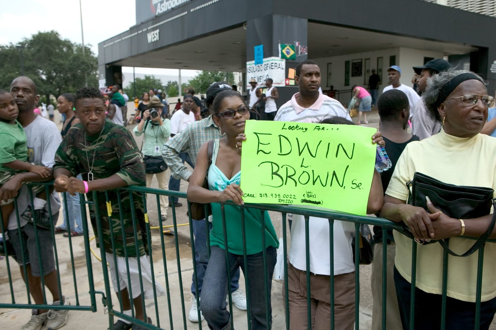 Pessoas apoiadas numa grade diante da entrada do estádio; uma mulher segura um cartaz verde escrito à mão procurando um parente