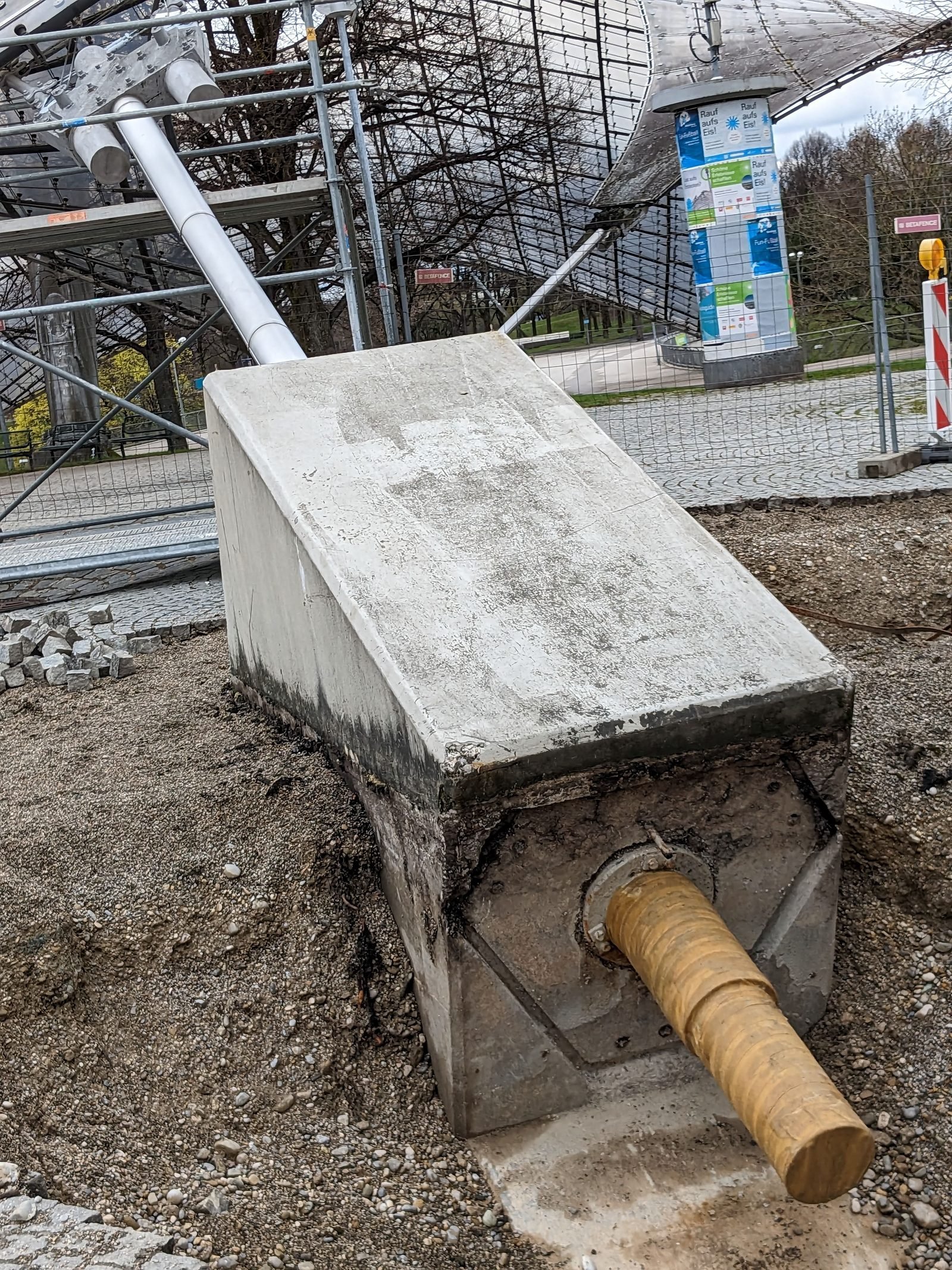 Bloco de concreto inclinado, parcialmente desenterrado, com um tubo grosso de onde sai um cabo; ao fundo, andaimes e a borda do teto