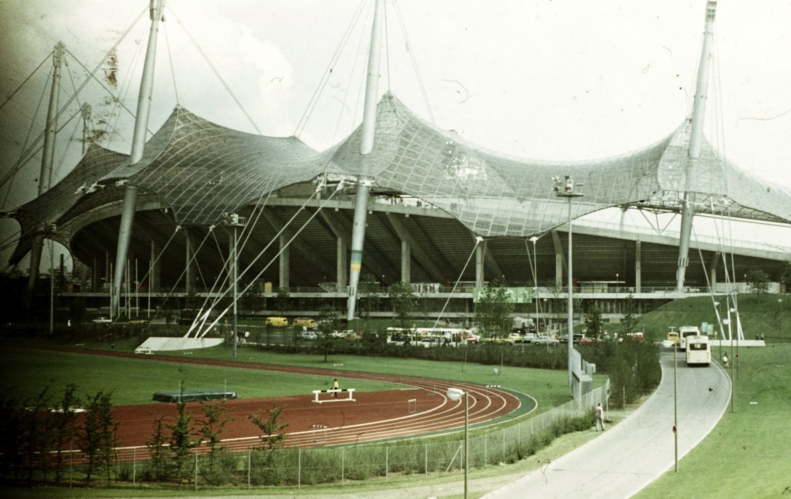 Color photograph from 1972: the stadium's transparent roof of cables and panels, hung from tall leaning masts, over a concrete grandstand; in the foreground, a red running track and a street with buses