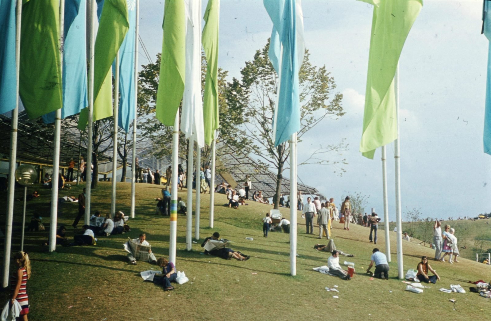 Fotografia colorida de 1972: pessoas sentadas e deitadas num gramado em declive, entre mastros com bandeiras verdes, azuis e brancas; à esquerda, a borda do teto de cabos