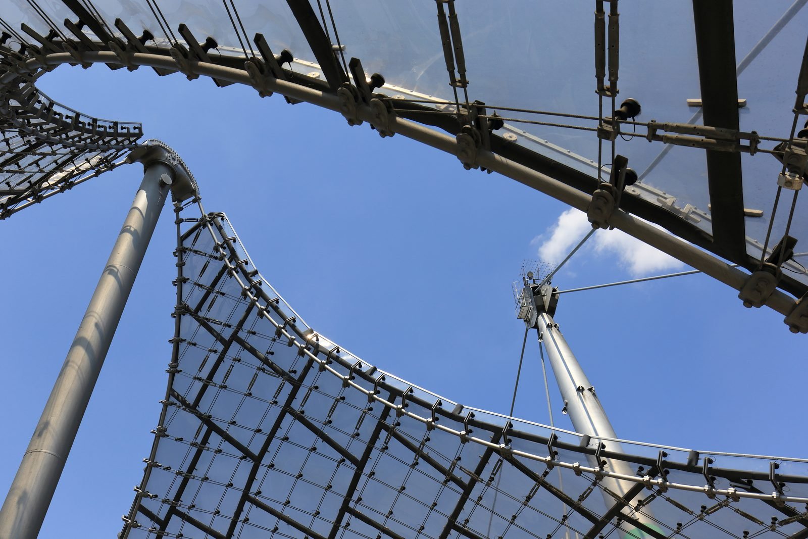 Vista de baixo para cima de uma borda do teto: a rede de cabos de aço em quadrados, as placas transparentes por cima, os grampos nos nós e um mastro cinza contra o céu azul