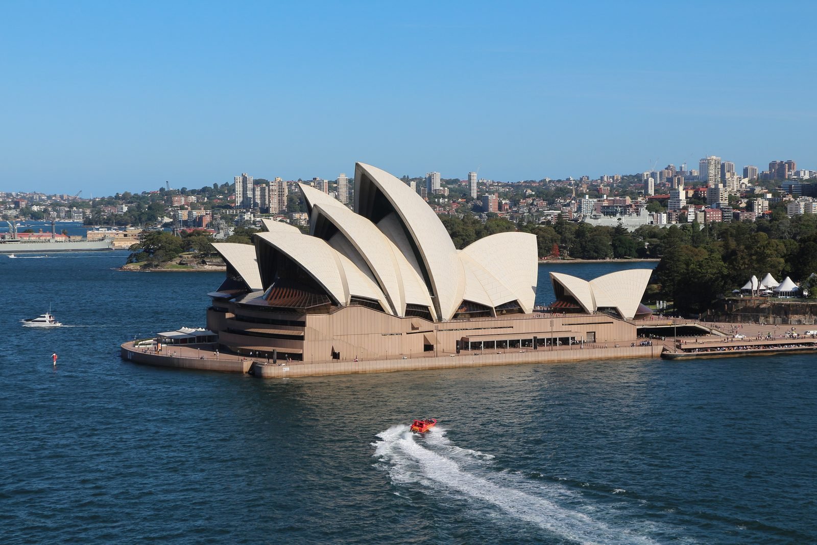 The Sydney Opera House seen from above by day: white shells on a sand-colored podium at the water's edge, blue water all around, a red boat leaving a trail of foam and the city in the background