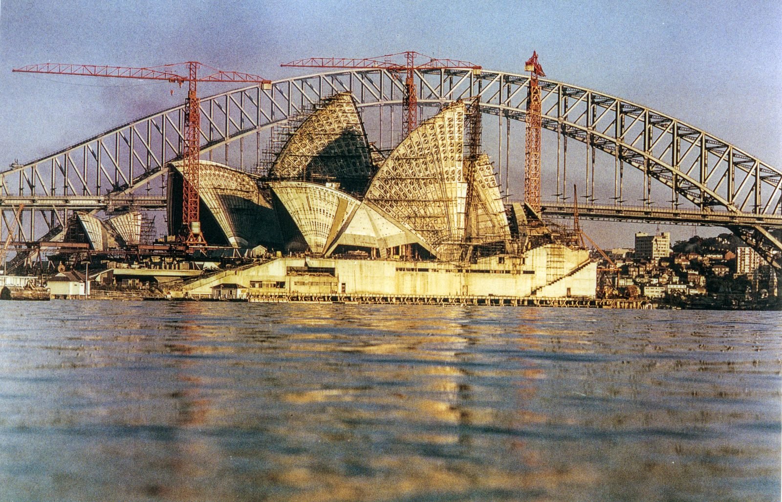 As conchas nervuradas ainda sem revestimento, com três gruas vermelhas, vistas do nível da água, e o arco de aço da Harbour Bridge atrás