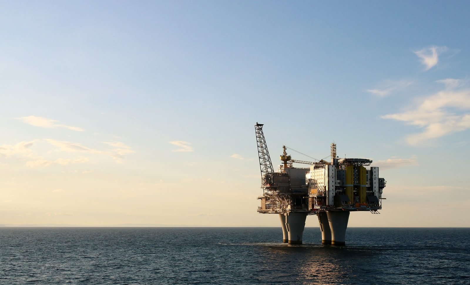 A gas platform on a calm sea at dusk, with four light-colored concrete columns rising from the water and a deck crowded with modules, a crane and a tower, to the right of a wide sky