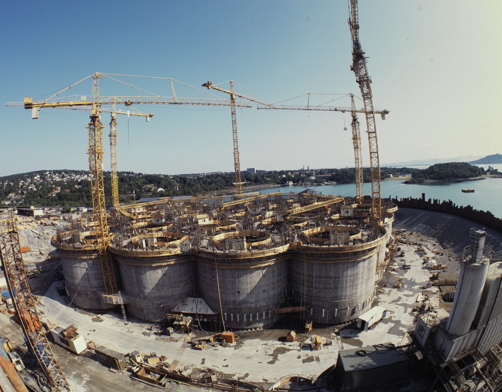 Vista gran angular de una obra a orillas de un fiordo: un grupo de grandes cilindros de concreto unidos, rodeados de grúas torre amarillas, con casas y mar al fondo
