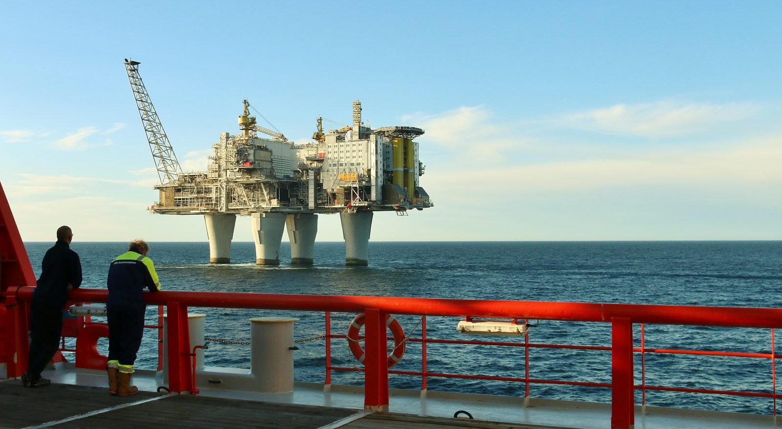 Dos hombres con overol apoyados en la baranda roja de un barco miran la plataforma en el mar, a lo lejos, con las cuatro columnas y la cubierta