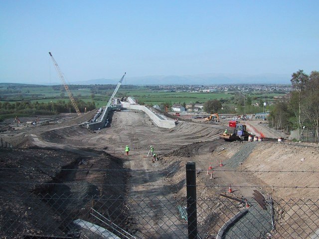 Obra vista desde lo alto: un largo lecho de tierra excavada entre muros de concreto, dos grúas de celosía y obreros con chaleco amarillo, con campos y montañas al fondo