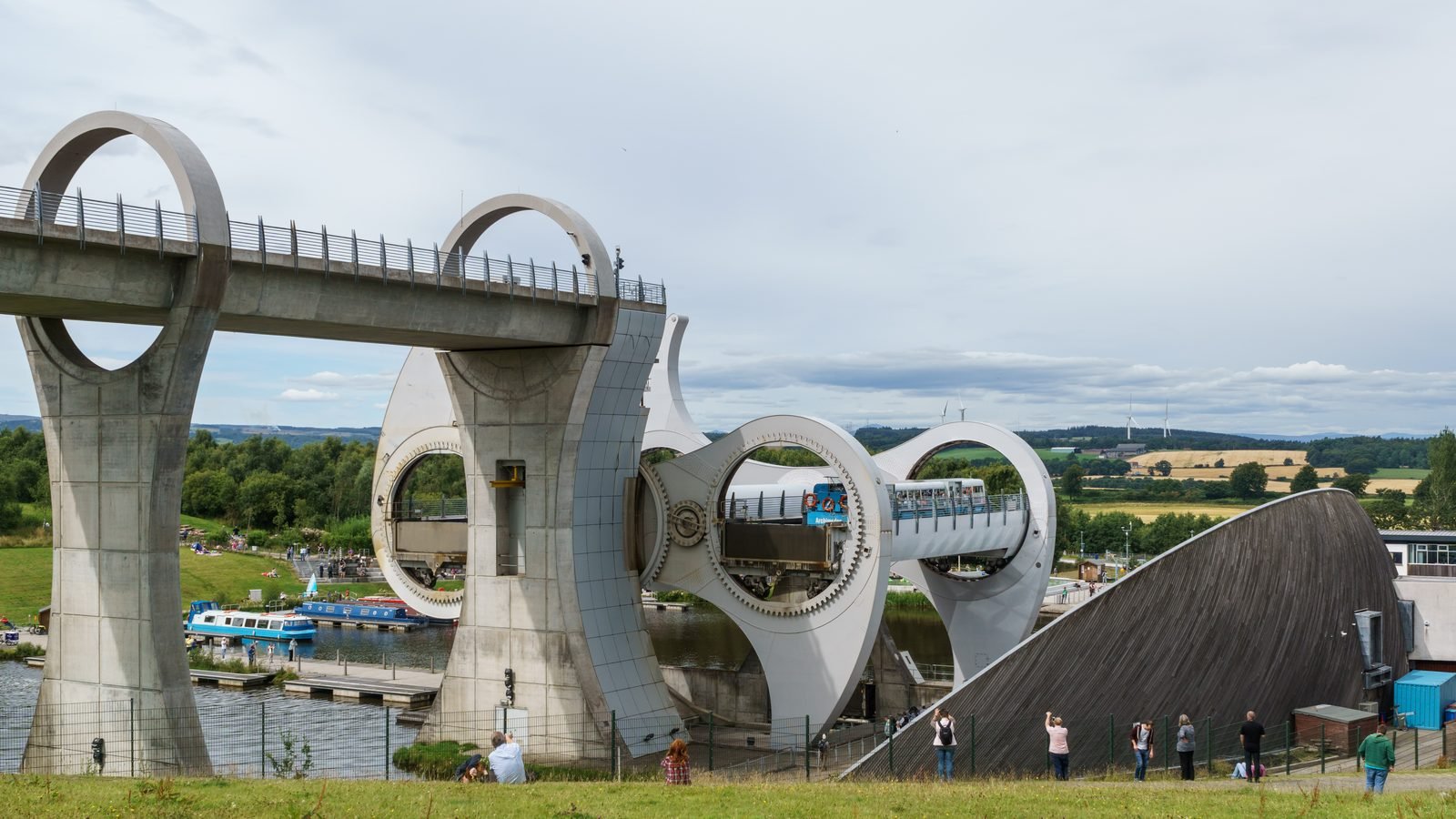 Large light-gray steel structure with two curved arms turning beside a concrete aqueduct carried on ring-shaped piers; a gondola with a boat is halfway up, with a sloping-roofed building on the right and people watching from a lawn
