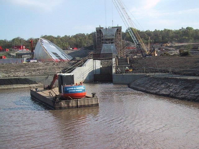 Dársena de agua turbia con una excavadora sobre una balsa; al fondo, un pilar de concreto envuelto en andamios, una grúa y la cáscara curva y vidriada de un edificio en construcción