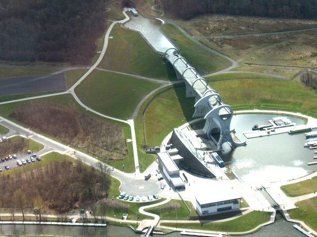 Vista aérea: un canal sale de un túnel entre árboles, sigue por un acueducto sobre pilares hasta la rueda, que baja a una dársena circular con barcos; al lado, el centro de visitantes y un estacionamiento