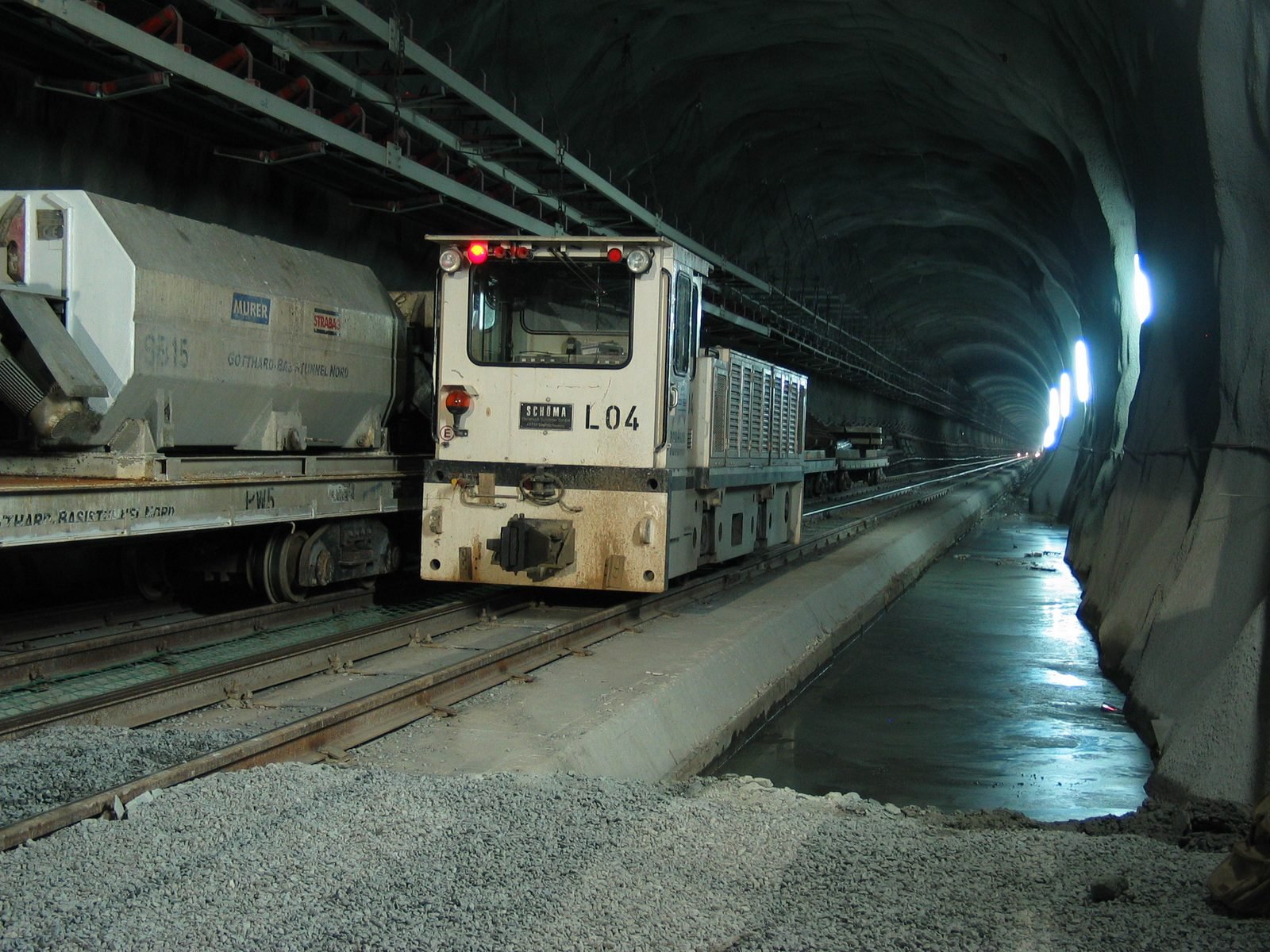 Interior of a dark rock tunnel under construction, with temporary rails, a small white service locomotive with its headlights on, a concrete car on the left and lights along the curved wall into the distance