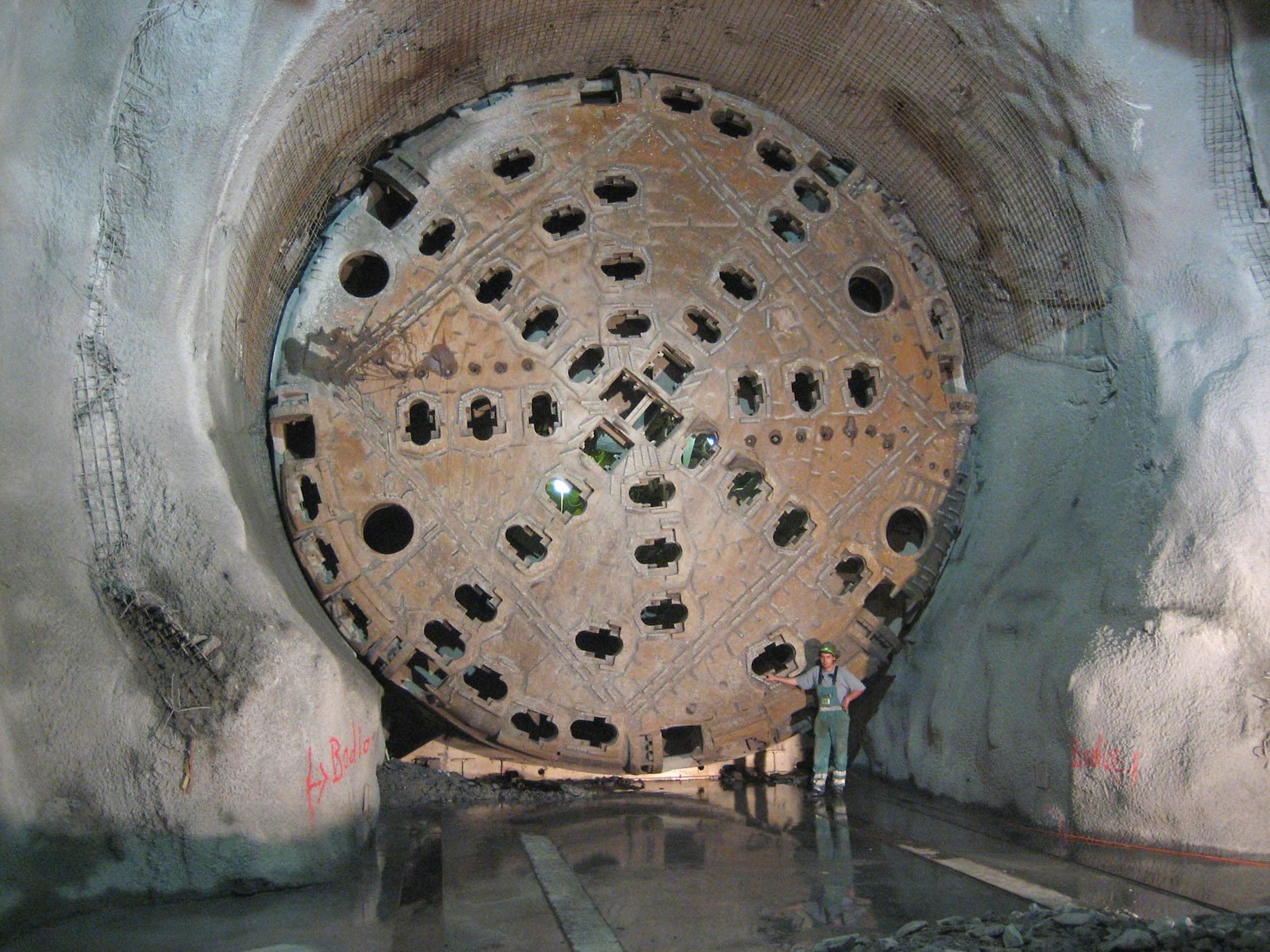 Inside a rock tunnel coated in shotcrete, the huge rust-colored circular cutterhead of a tunnel boring machine, full of openings; a worker in a helmet and coveralls stands beside it, reaching about a quarter of the way up the disc