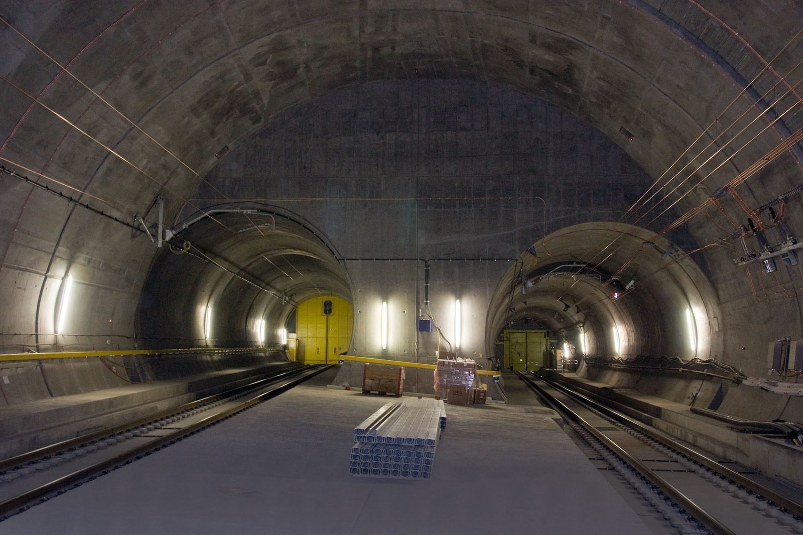 Large arched concrete cavern, with two tunnel mouths side by side at the back, each closed by a yellow gate, rails on the floor and construction materials stacked in the middle