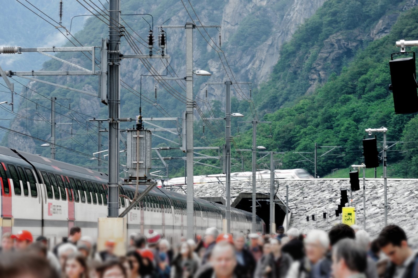 Blurred crowd in the foreground at an open-air station, a double-deck train standing on the left and, in the background, between overhead-line masts and green mountains, the tunnel mouth with a train coming out