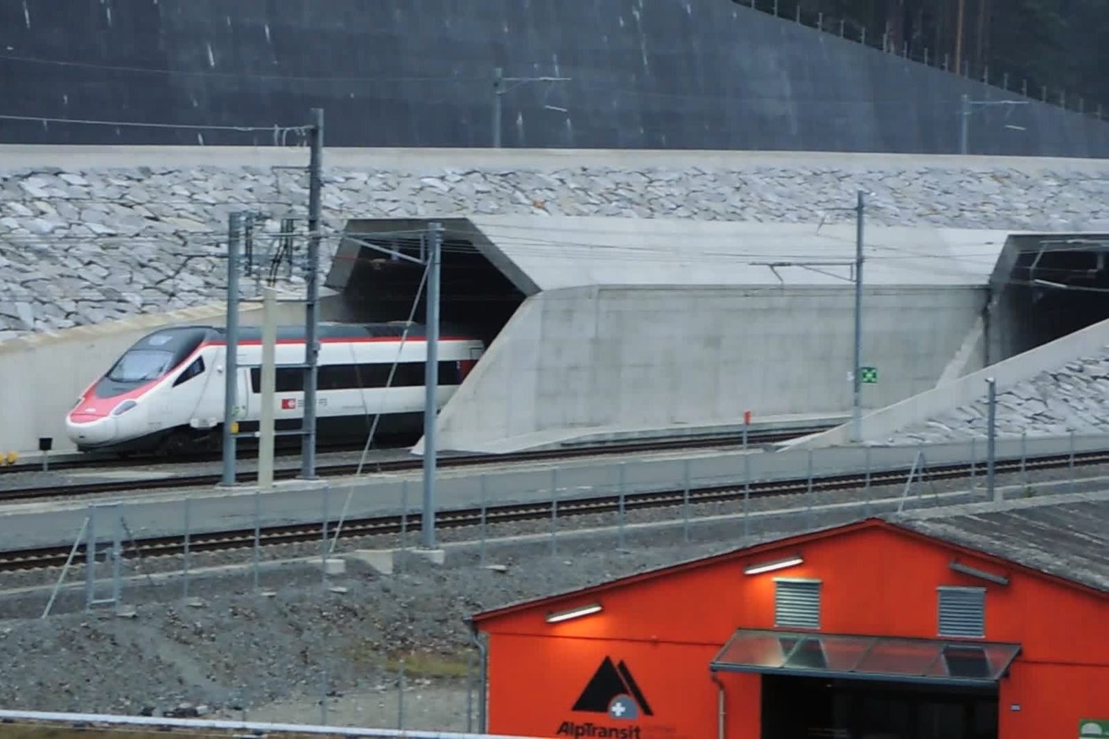 White pointed-nose train coming out of one of two smooth concrete tunnel mouths, beneath a rocky embankment; in the foreground, rails and the roof of a red shed