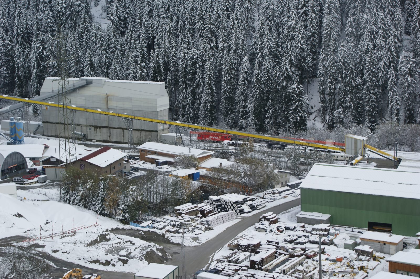 View from above of a snow-covered construction site in a valley of pine trees, with sheds, a tall gray building, stacks of pipes and a long yellow conveyor belt crossing the scene